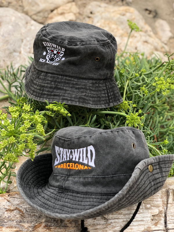 Two hats displayed on wood and greenery: the Stay Wild bucket hat with front panther logo, and the Stay Wild cowboy hat with “STAY WILD BARCELONA” embroidered on the back in white and yellow