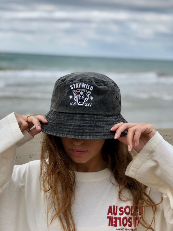 Woman wearing the Stay Wild bucket hat at the beach, holding the brim down over her face with ocean in the background