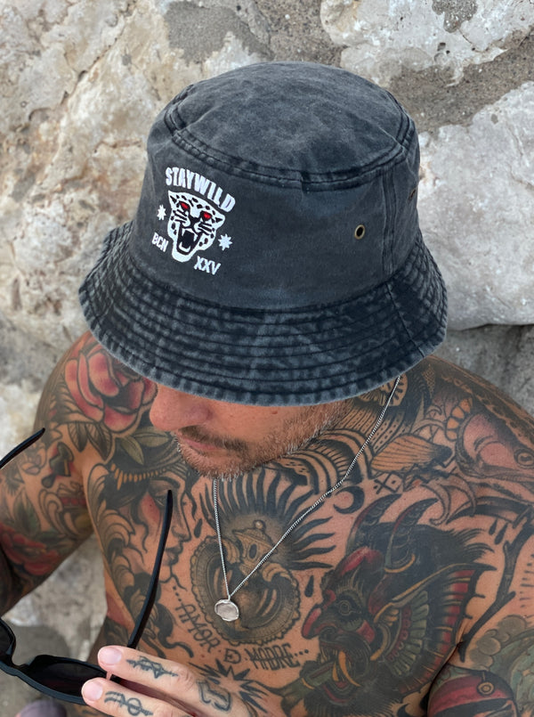 Man wearing the black Stay Wild bucket hat with white panther logo and “BCN XXV” text, sitting shirtless against a stone wall
