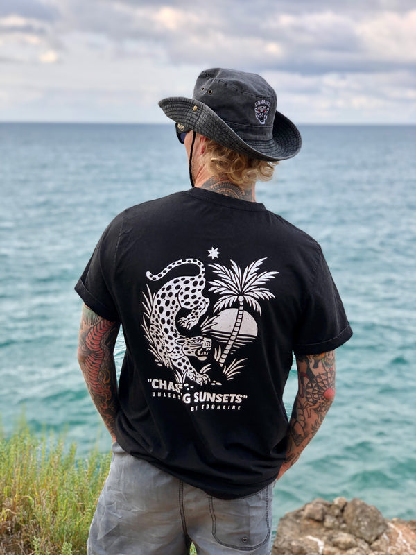 Person wearing the black Chasing Sunsets t-shirt facing the sea, with a bold leopard and palm tree design on the back