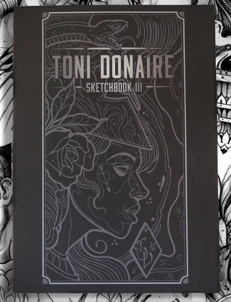 Black sketchbook with metallic silver print on the cover. The design shows a woman’s profile with floral and ornamental motifs, framed by fine linework
