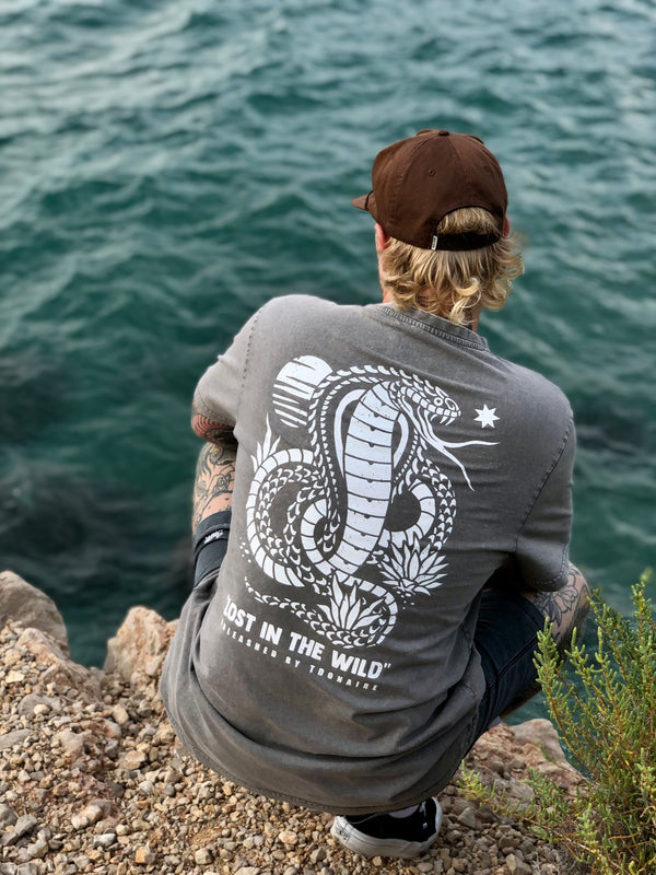 Man sitting on rocks by the sea wearing the Lost in the Wild tee, showing the large back print of a snake and agave plants
