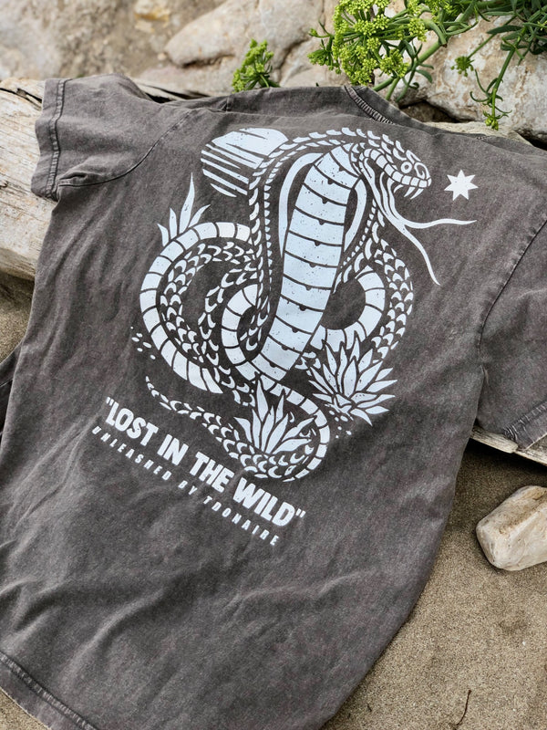 Faded black t-shirt with white snake and desert plant design laid flat on sand and wood, featuring the text “Lost in the Wild”