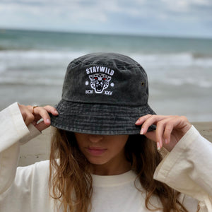 Woman wearing the Stay Wild bucket hat at the beach, holding the brim down over her face with ocean in the background