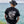 Person wearing the black Chasing Sunsets t-shirt facing the sea, with a bold leopard and palm tree design on the back