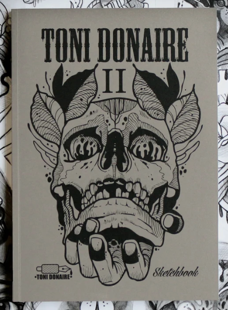 Close-up of the Sketchbook II cover by Toni Donaire, featuring a bold black ink skull illustration with symmetrical elements on a beige background.