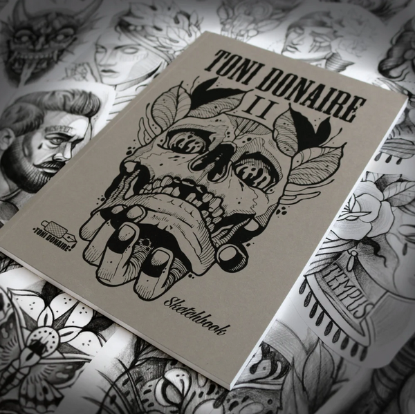 Angled view of the closed sketchbook lying on a surface full of pencil drawings, highlighting its textured cover and bold black linework.