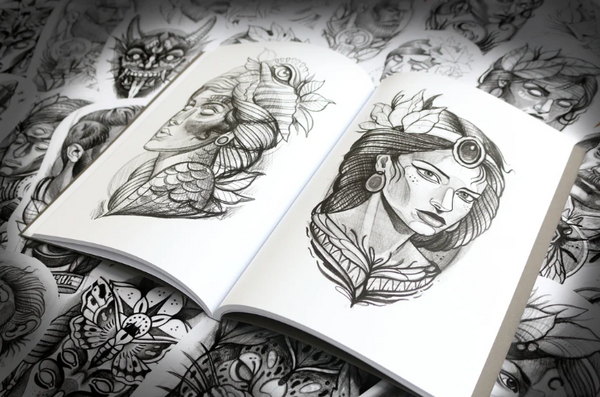 Open sketchbook displaying two pencil portraits of women, both in classic tattoo flash style. One has fish scales and a fin on her head; the other wears a jewel on her forehead and earrings. Surrounded by other sketch pages underneath.